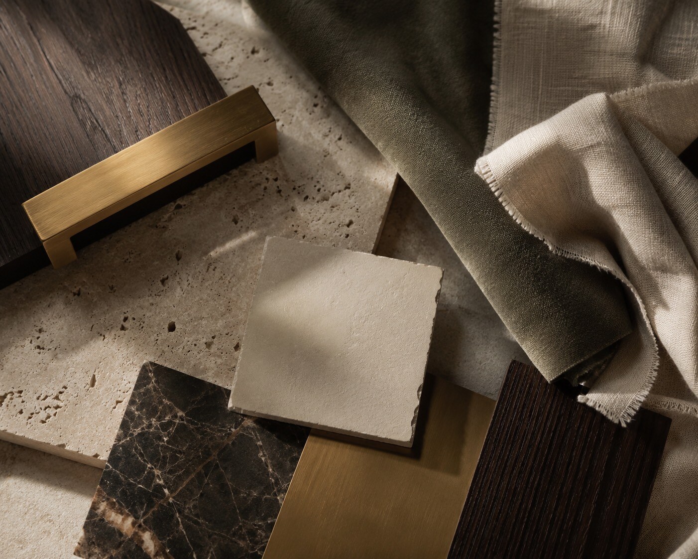 A materials layout in raking light — fumed oak, travertine, brushed brass, dark marble, honed limestone, olive velvet, and raw-edged linen